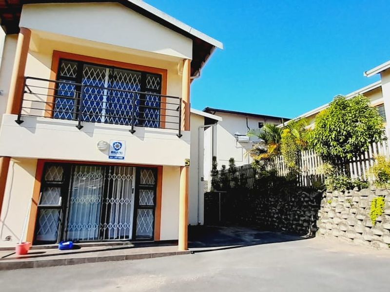 Front exterior view of a two-story cluster unit featuring a balcony with security gates, paved driveway, and a stone retaining wall.