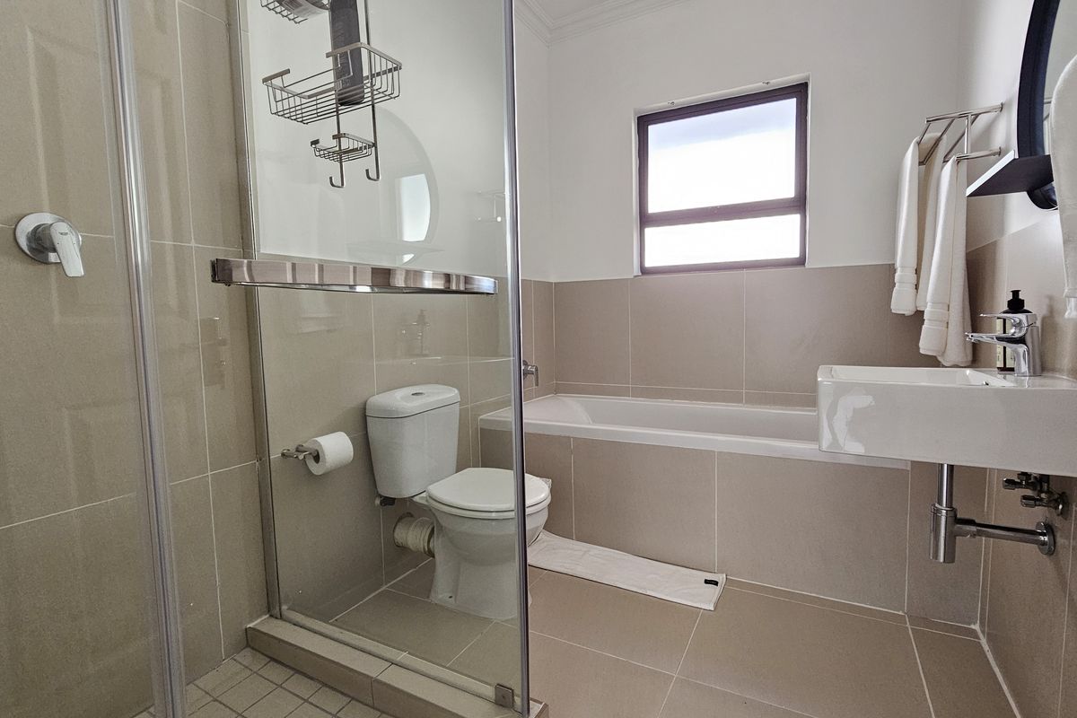 A modern bathroom featuring a glass-enclosed shower, a built-in bathtub, a wall-mounted basin, and neutral floor-to-ceiling tiling.
