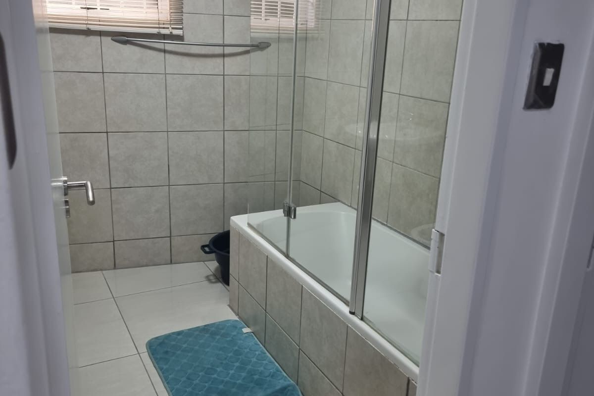 A functional bathroom featuring a tiled bathtub with a glass shower screen, neutral wall and floor tiles, and a window with fitted blinds.