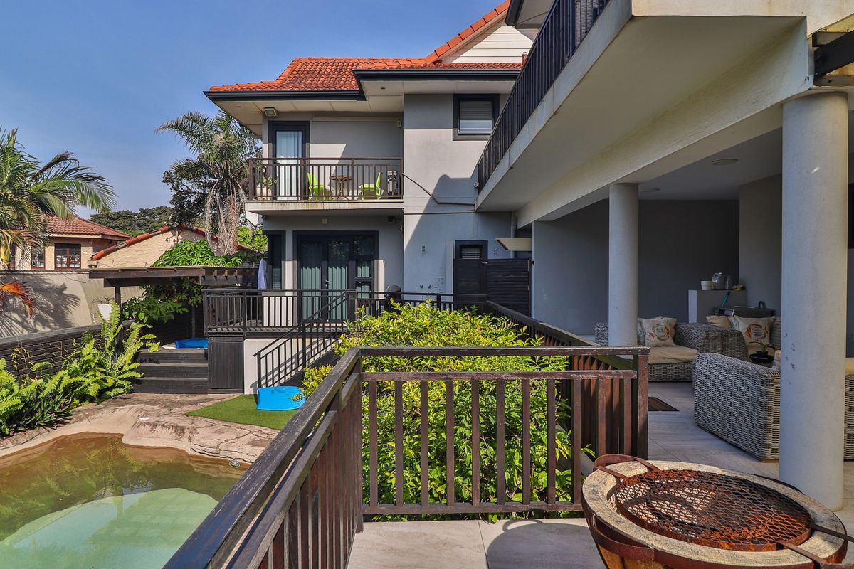 Rear exterior view featuring a covered patio, balcony, and swimming pool area with established garden landscaping and tiled outdoor flooring.
