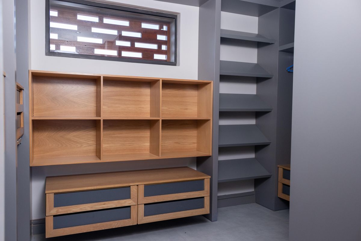 A modern walk-in closet featuring custom built-in cabinetry with a mix of light wood shelving and dark grey drawers, complemented by a decorative breeze block window.