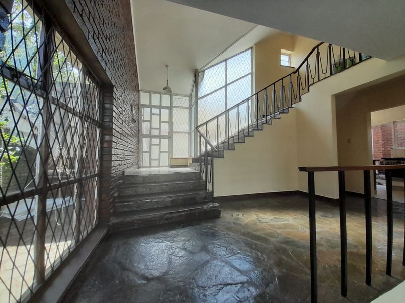 Spacious entrance hall featuring exposed brickwork, stone flooring, and a prominent staircase with wrought iron railing, illuminated by large windows.