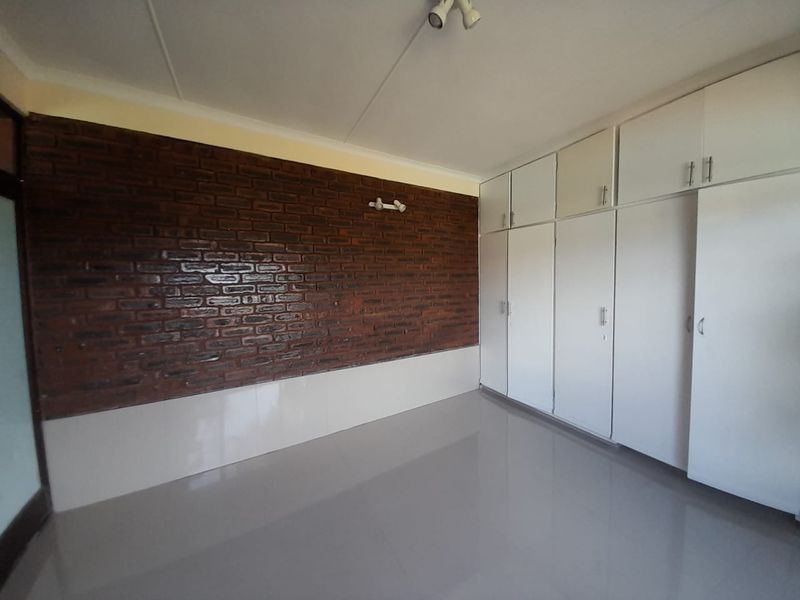 A spacious bedroom featuring a distinctive exposed brick feature wall, neutral tiled flooring, and extensive built-in cupboard storage.