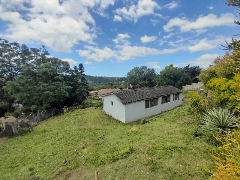 A rear view of a detached outbuilding or townhouse structure set within a large, sloped garden area with mature trees and boundary fencing.
