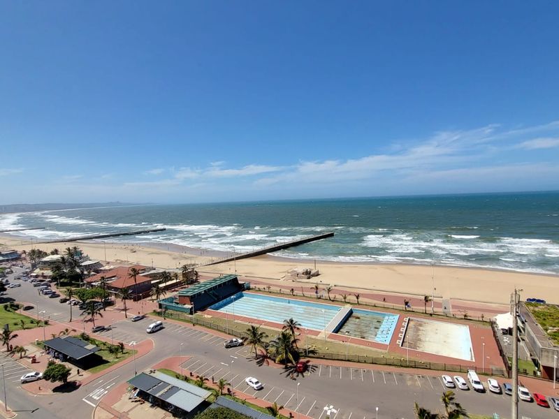 A high-angle panoramic view overlooking the coastline, beach, and public swimming pool facilities.