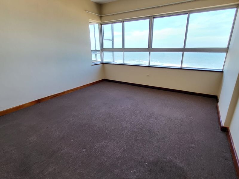 An empty bedroom featuring large windows with ocean views, carpeted flooring, and timber skirting boards, offering a blank canvas for renovation.