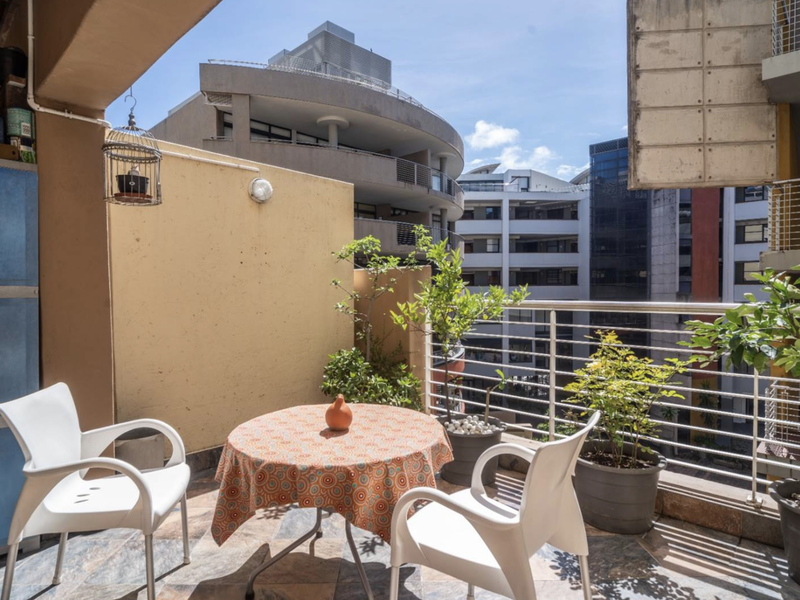 A covered balcony featuring tiled flooring, a metal safety balustrade, and built-in storage, offering an urban view of the surrounding apartment complex.