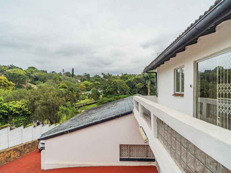 Rear exterior view of the property featuring a tiled roof, glass block wall detailing, and a paved driveway overlooking lush greenery.