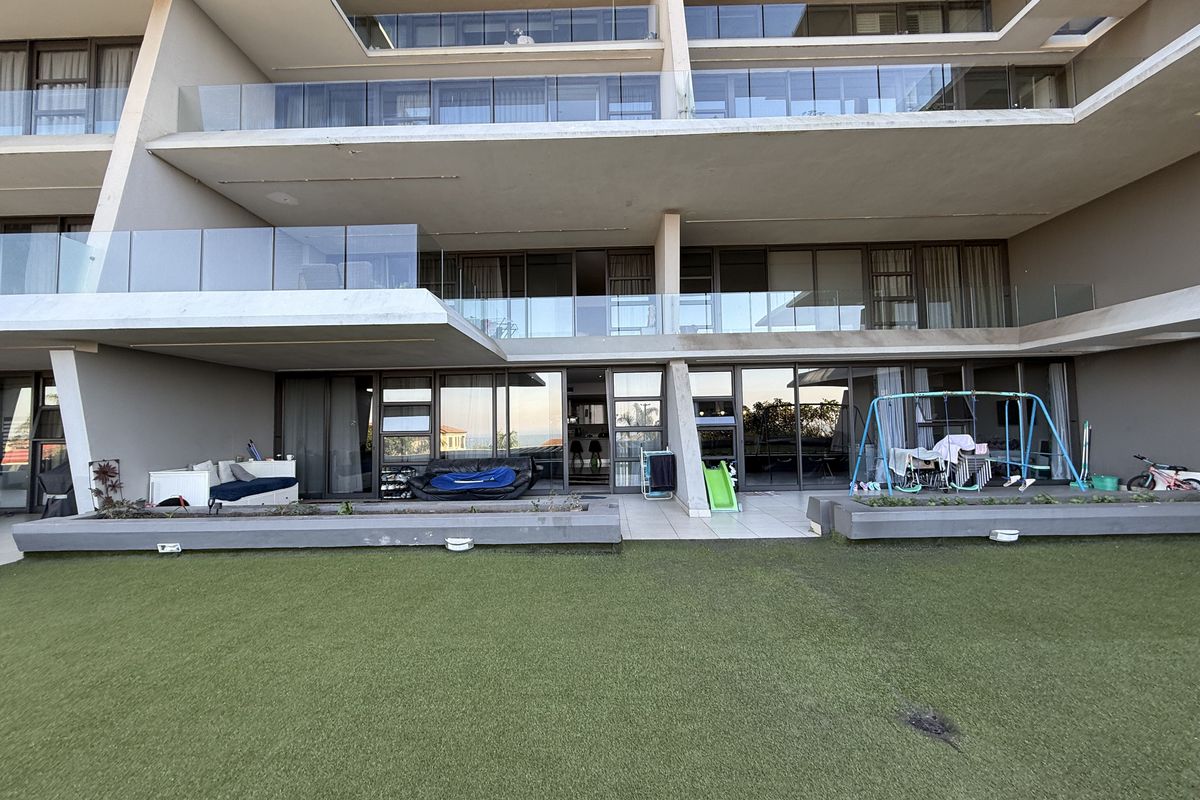 Rear view of a modern apartment building featuring multiple levels of balconies with glass balustrades and ground floor access to an artificial lawn area.