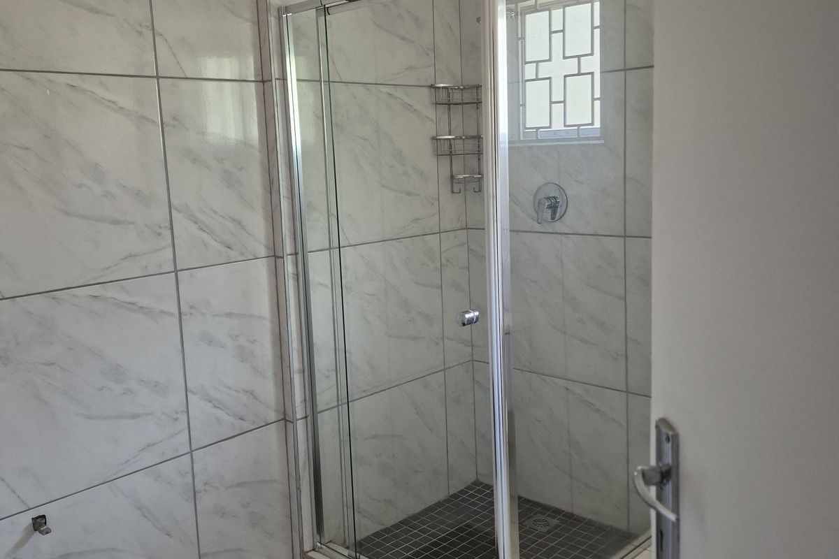 A clean bathroom featuring a corner shower enclosure with glass doors, marble-look wall tiles, and a small window with security bars.