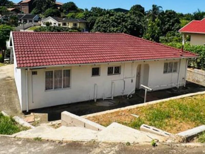 Rear view of a single-story residential house featuring a tiled roof, concrete driveway, and tiered retaining walls in a suburban setting.