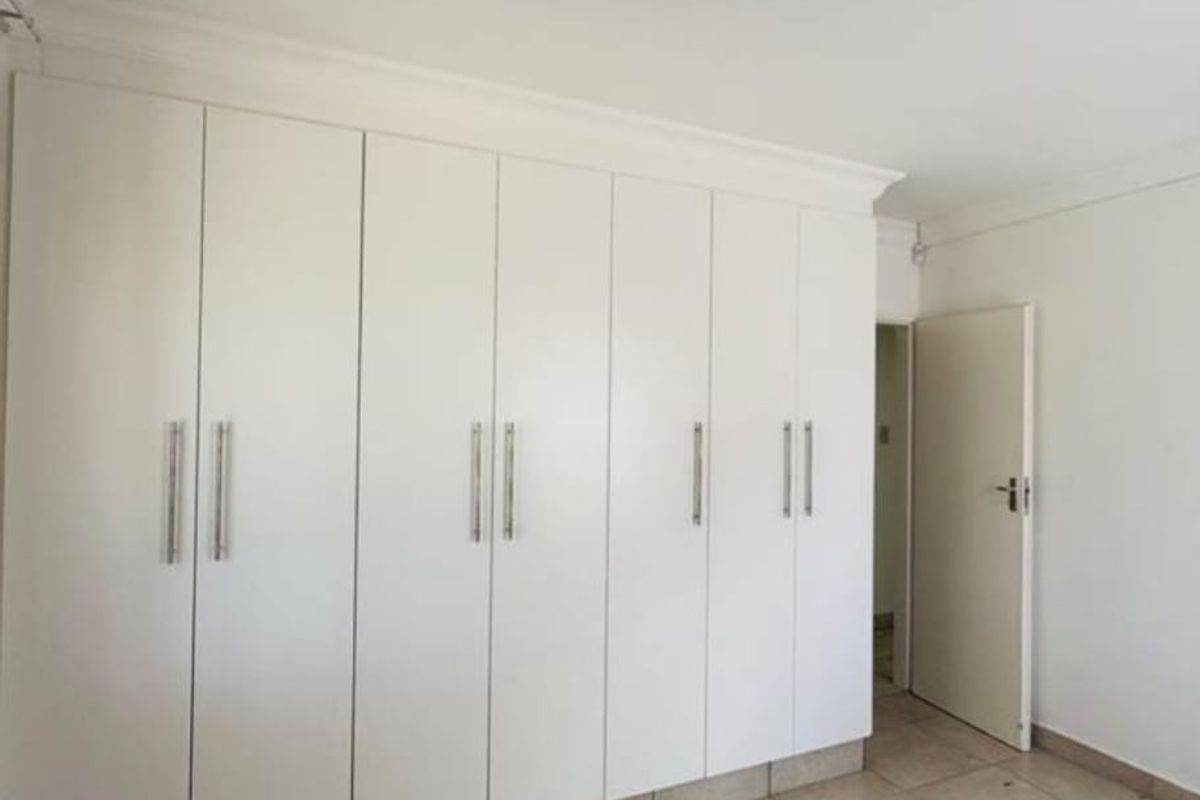 A clean, empty bedroom featuring extensive white built-in cupboards, neutral tiled flooring, and simple cornice detailing.