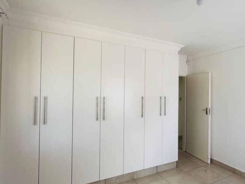 A clean, empty bedroom featuring extensive white built-in cupboards, neutral tiled flooring, and simple cornice detailing.