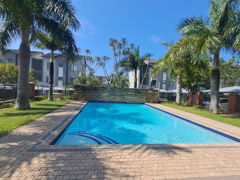 A well-maintained communal swimming pool area featuring a stone-clad water feature, paved surround, and landscaped garden setting within an apartment complex.
