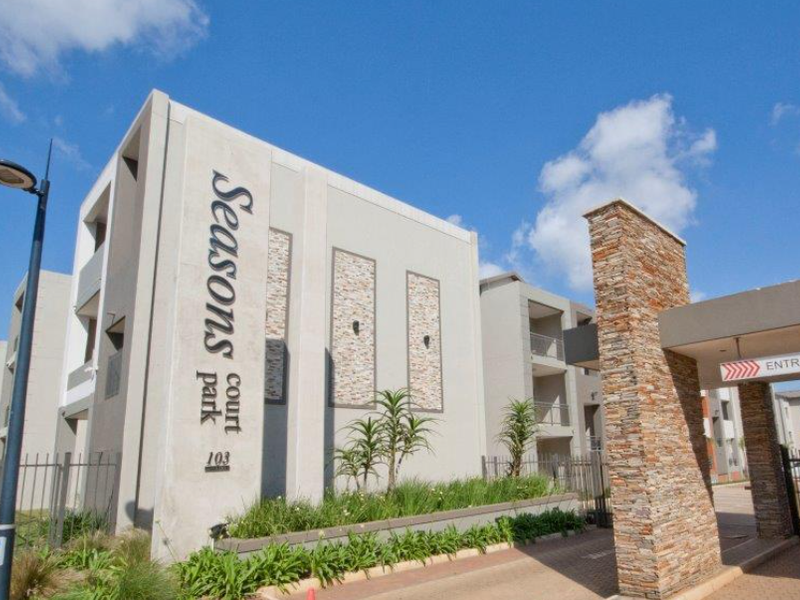 Exterior view of the Seasons Court Park apartment complex entrance, featuring modern stone cladding, secure gated access, and landscaped garden beds.