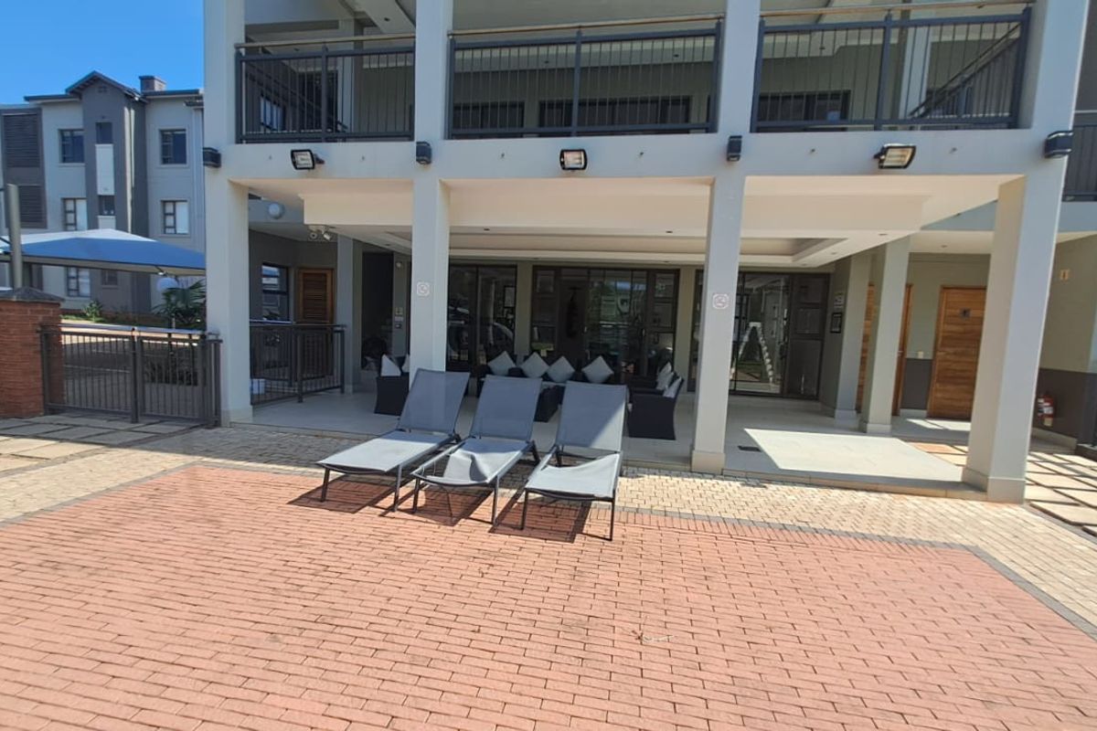 A spacious covered patio area featuring paved flooring, structural pillars, and integrated exterior lighting, providing a seamless transition to the outdoor pool area.