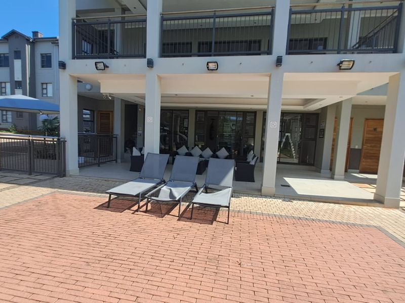 A spacious covered patio area featuring paved flooring, structural pillars, and integrated exterior lighting, providing a seamless transition to the outdoor pool area.