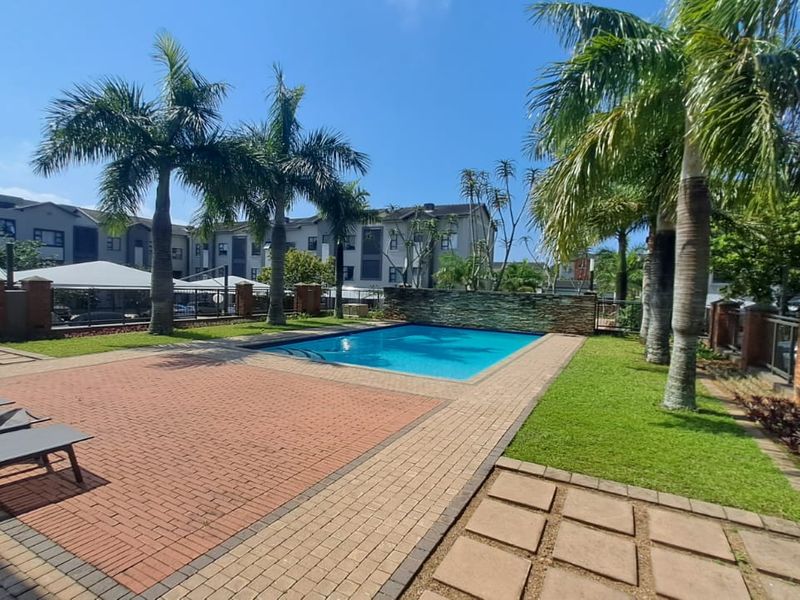 Communal swimming pool area featuring paved decking, landscaped lawn, and established palm trees within a residential apartment complex.