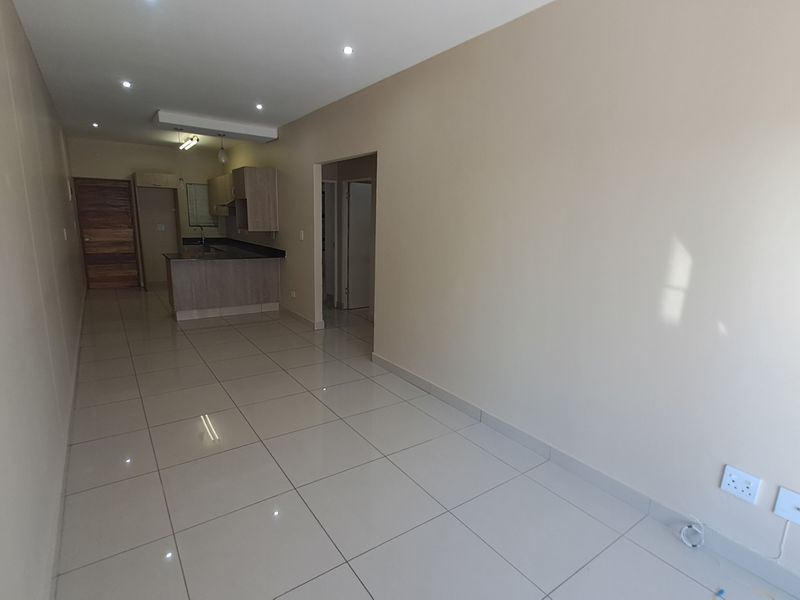 An open-plan living and kitchen area featuring neutral tiled flooring, recessed lighting, and modern kitchen cabinetry in a clean, vacant space.