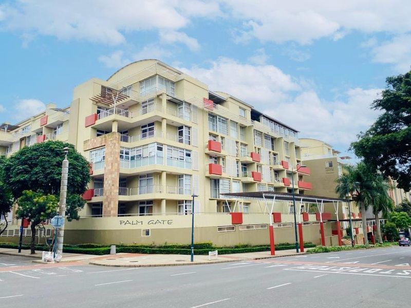A street-level view of the Palm Gate apartment complex, showcasing its multi-story facade, balconies, and surrounding perimeter infrastructure.