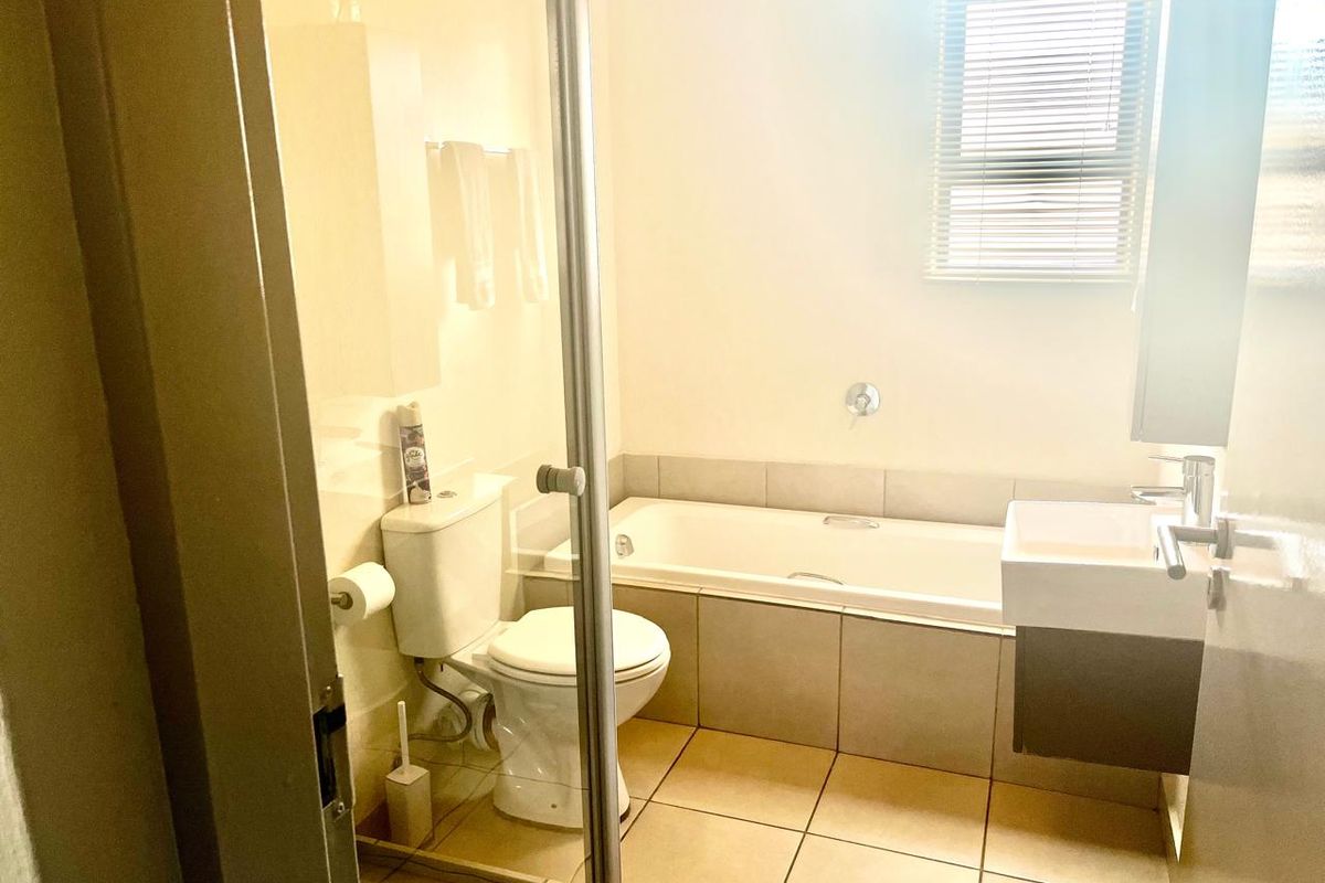 A functional bathroom featuring a built-in bathtub, a wall-mounted vanity basin, a toilet, and tiled flooring with natural light from a window.