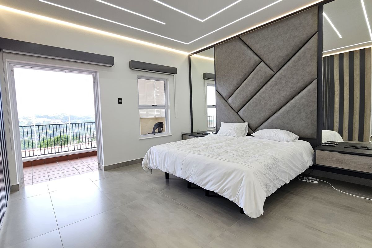 Modern master bedroom featuring a large upholstered headboard, recessed LED ceiling lighting, tiled flooring, and direct access to a private balcony with scenic views.