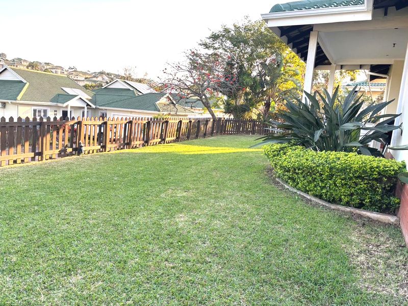 A well-maintained private garden featuring a lush lawn, landscaped garden beds, and a timber picket fence, adjacent to a covered patio area.