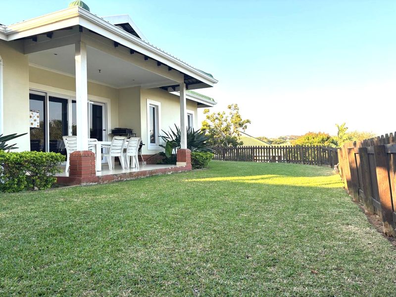 A well-maintained rear exterior view featuring a covered patio with brick pillars, sliding glass doors, and a spacious lawn enclosed by a timber fence.
