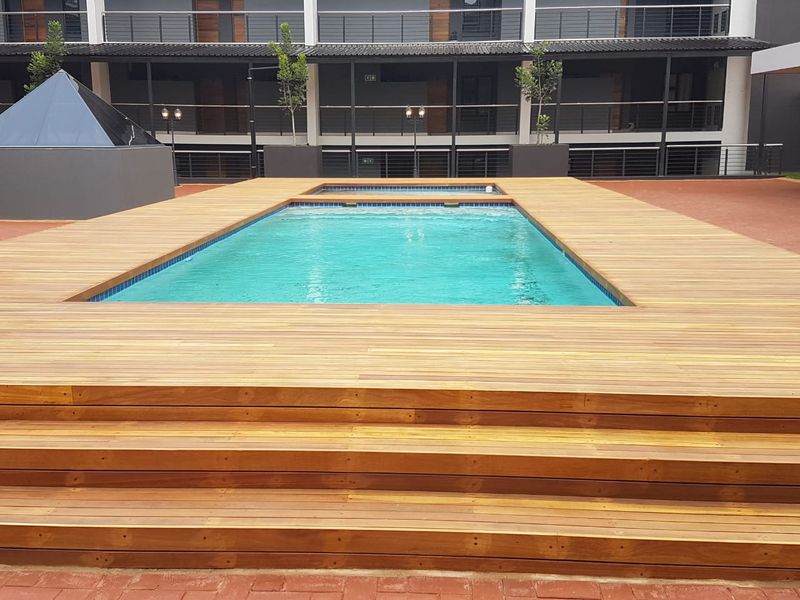 A well-maintained swimming pool area featuring extensive timber decking, integrated steps, and modern apartment complex surroundings.