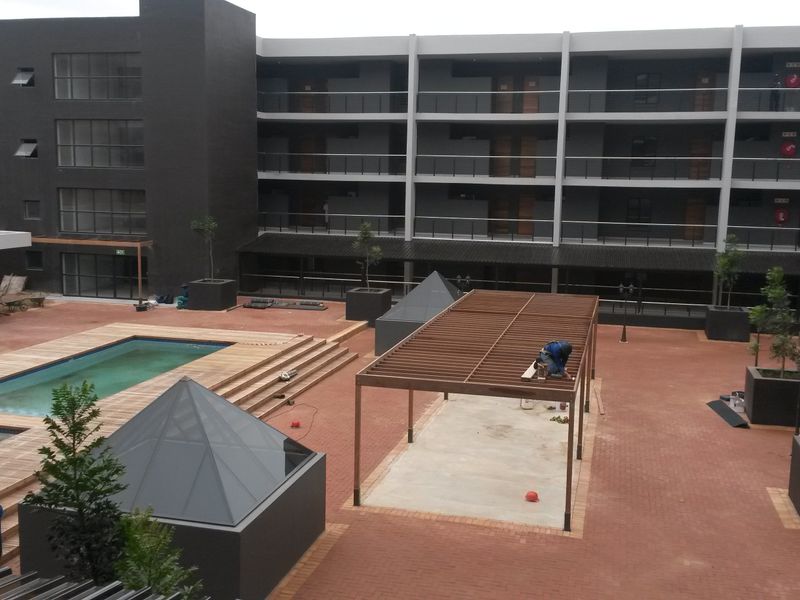 A modern apartment complex courtyard featuring a swimming pool, timber decking, and a central pergola structure within a paved outdoor area.