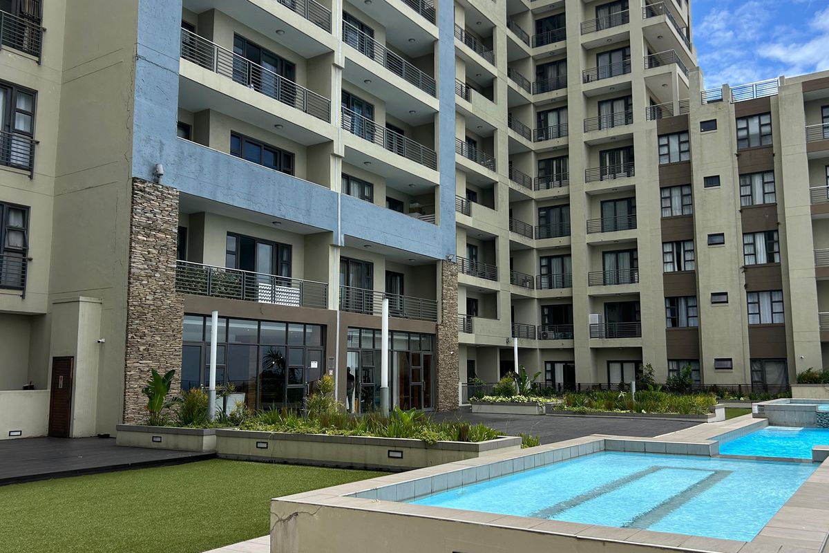 Exterior view of a modern apartment complex featuring multiple balconies, stone facade accents, a swimming pool, and landscaped communal outdoor areas.