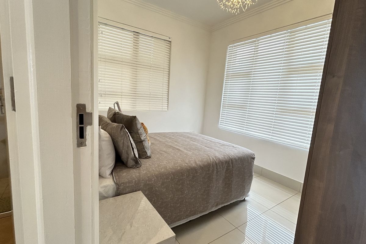 A bright bedroom featuring tiled flooring, dual windows with venetian blinds, and a modern light fixture, with an adjacent view into a bathroom.
