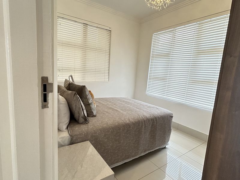 A bright bedroom featuring tiled flooring, dual windows with venetian blinds, and a modern light fixture, with an adjacent view into a bathroom.