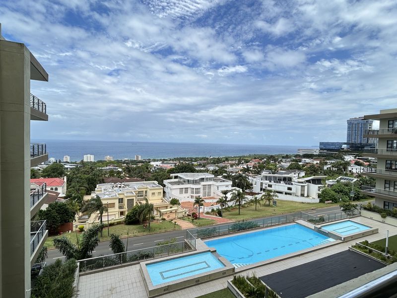 Elevated panoramic view overlooking the complex swimming pool, surrounding residential neighborhood, and the distant ocean coastline.