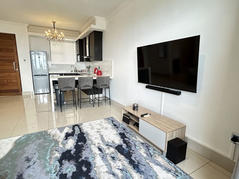 Modern open-plan living area featuring a sleek kitchen with island seating, tiled floors, and a wall-mounted television, creating a functional and contemporary living space.