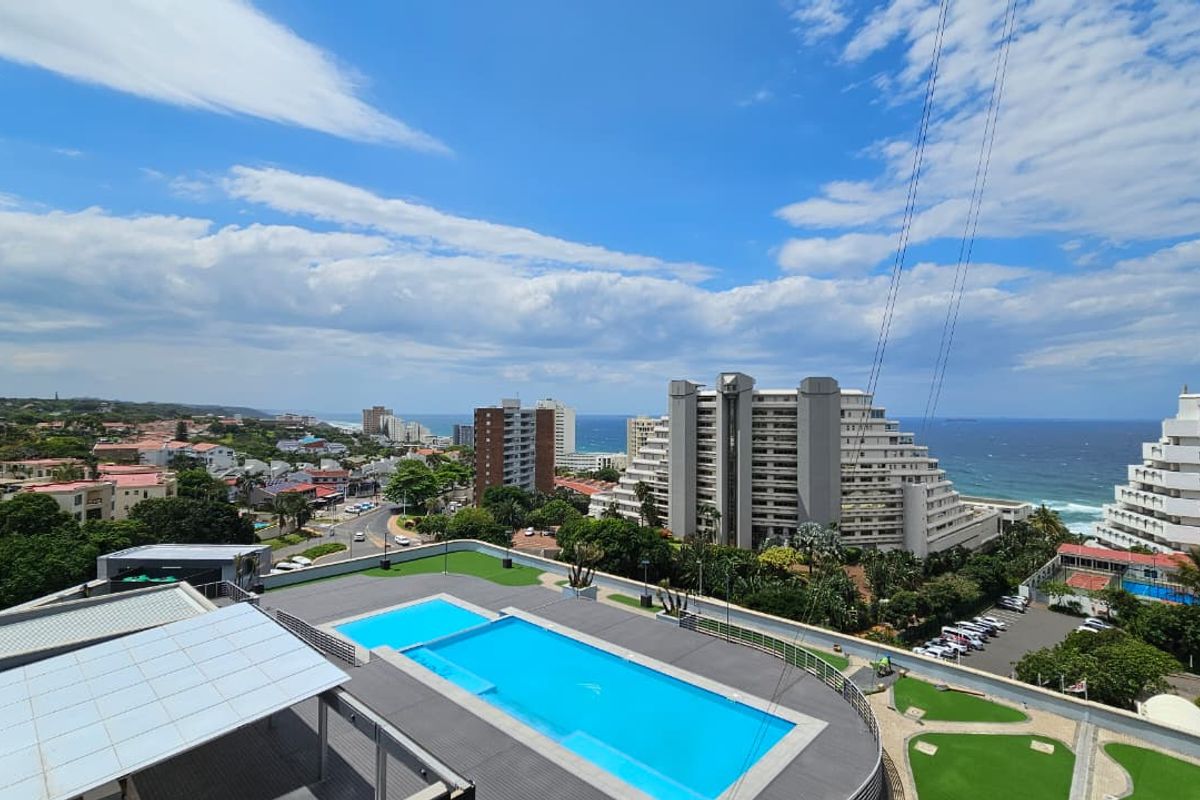 Elevated aerial view showcasing the apartment complex pool area, surrounding coastal landscape, and ocean horizon.