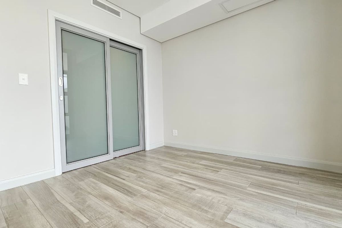 A bright, empty bedroom featuring modern wood-look tile flooring, neutral walls, and a sliding glass door providing access to the exterior.