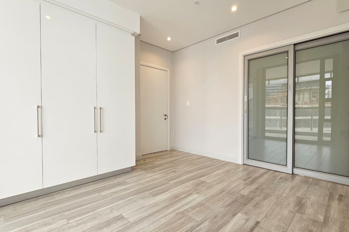 A bright, modern bedroom featuring built-in white cabinetry, wood-look flooring, recessed lighting, and sliding glass doors leading to a private balcony.