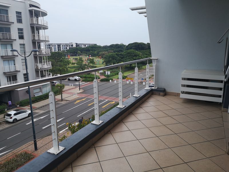 A spacious balcony featuring tiled flooring and a modern stainless steel balustrade, offering an elevated view of the surrounding street and park area.