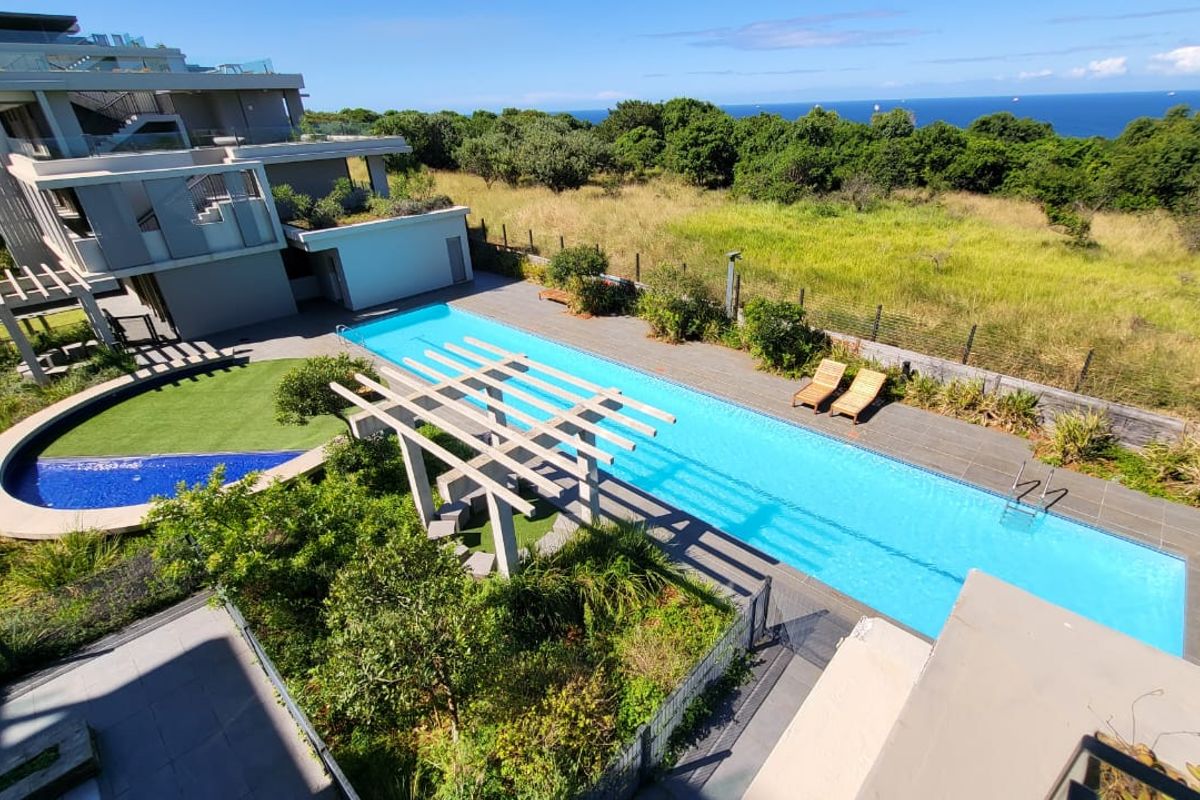 An elevated view of a modern apartment complex featuring a long lap pool, timber pergola, and landscaped garden area with ocean views.