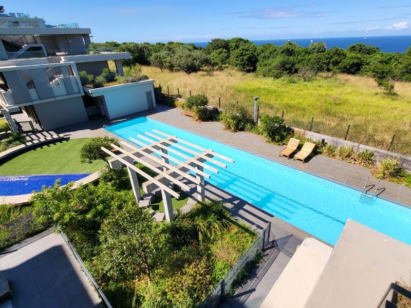 An elevated view of a modern apartment complex featuring a long lap pool, timber pergola, and landscaped garden area with ocean views.
