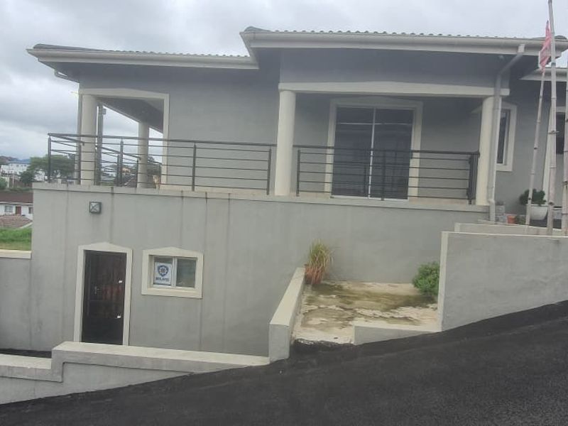 Front exterior view of a multi-level house featuring a paved driveway, elevated balcony with metal railings, and secure gated entrance.
