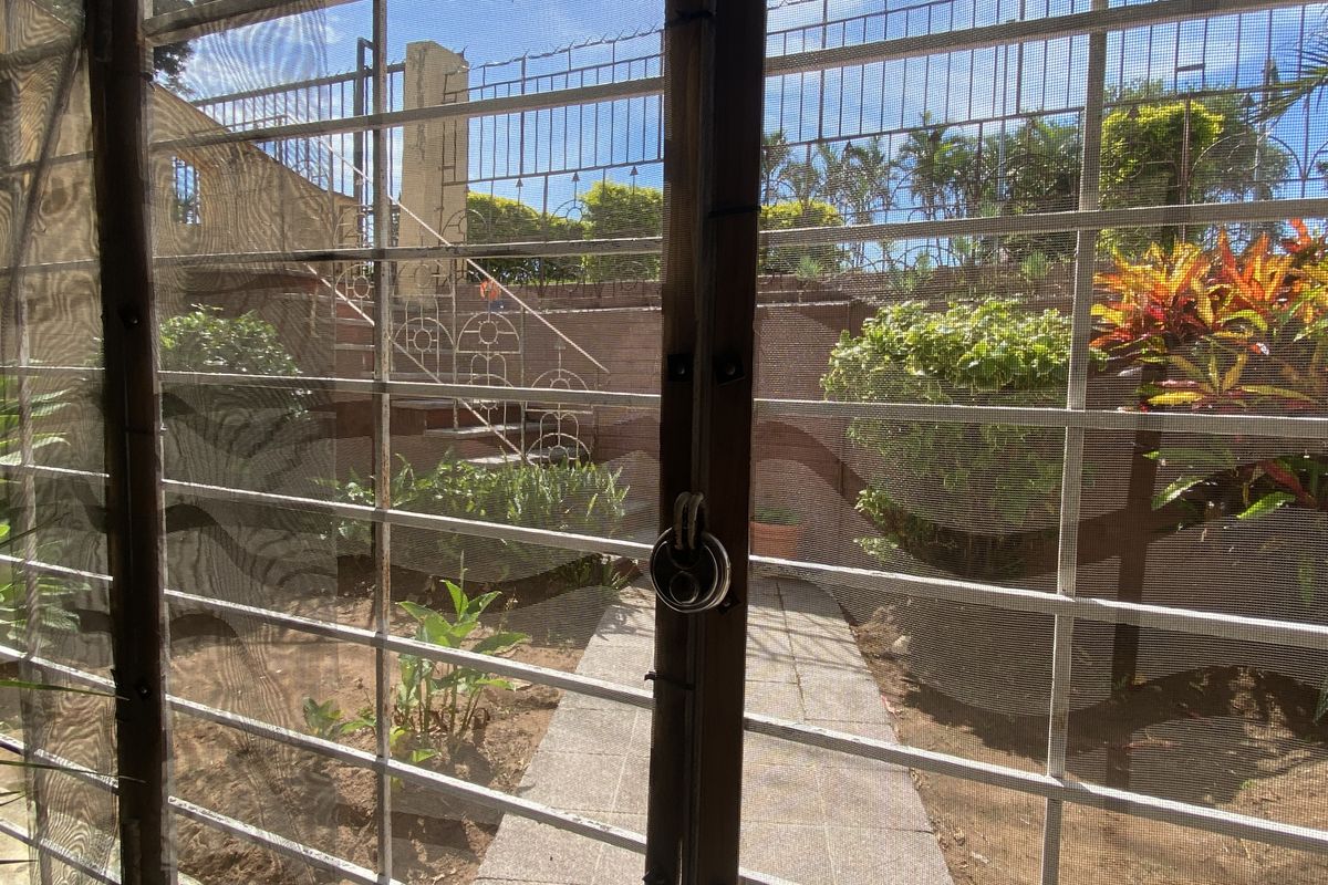 View of a rear garden area featuring a paved walkway, perimeter security fencing, and established landscaping, viewed through a security gate.