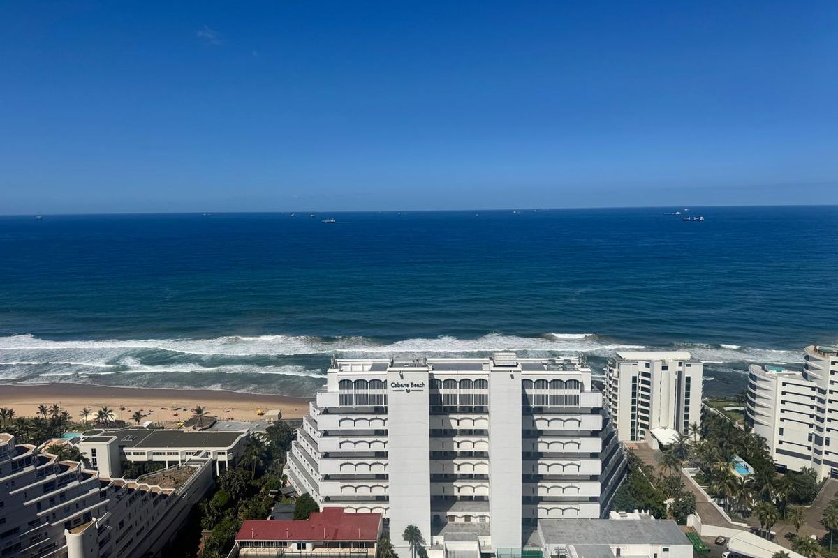 Elevated aerial view overlooking the coastline, featuring the ocean, sandy beach, and surrounding apartment complexes.