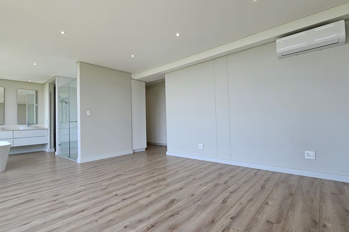 Spacious bedroom featuring modern laminate flooring, recessed ceiling lighting, a wall-mounted air conditioning unit, and an integrated en-suite bathroom with a contemporary vanity and shower.