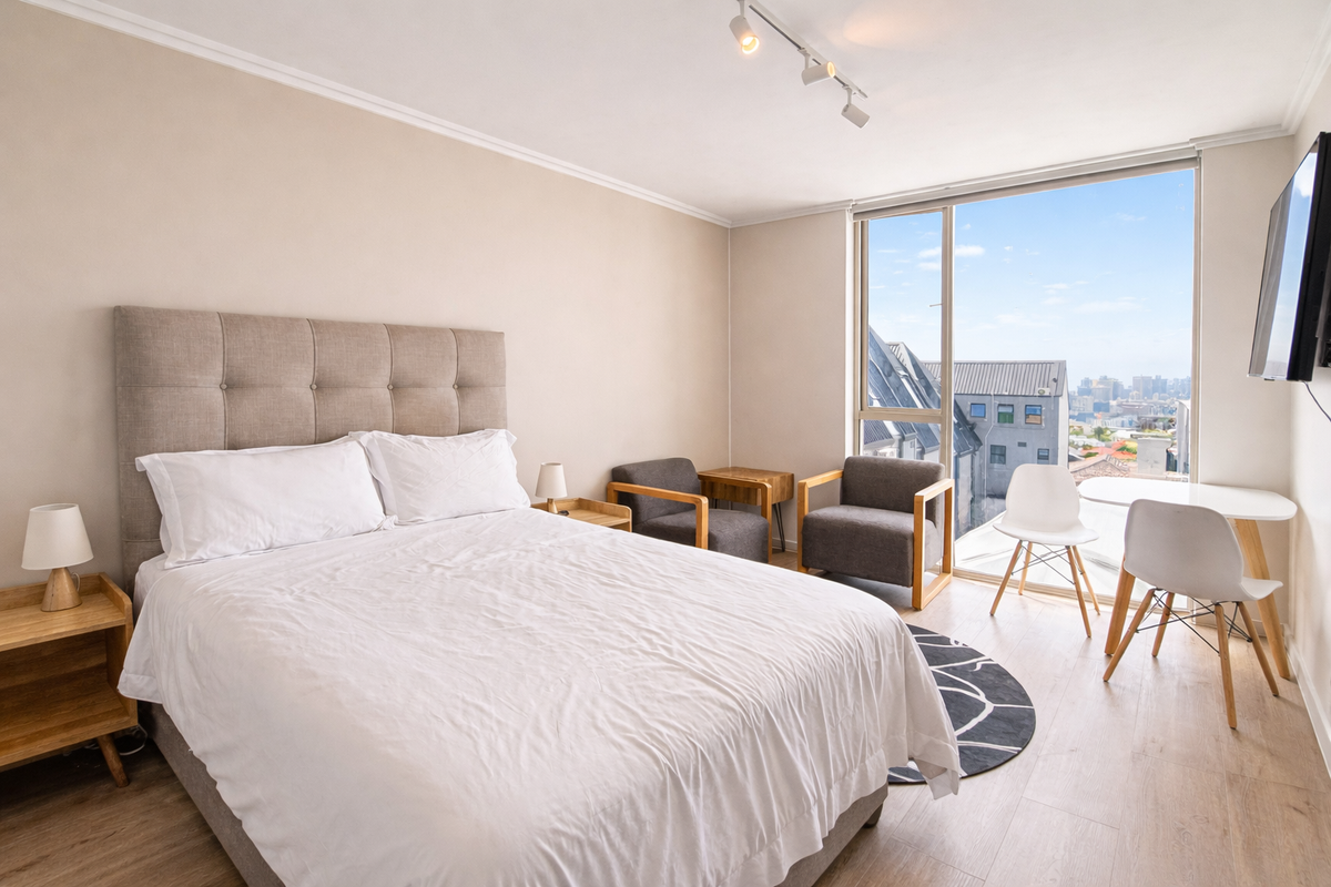 A bright bedroom featuring light wood-look flooring, a large window with city views, and modern track lighting.