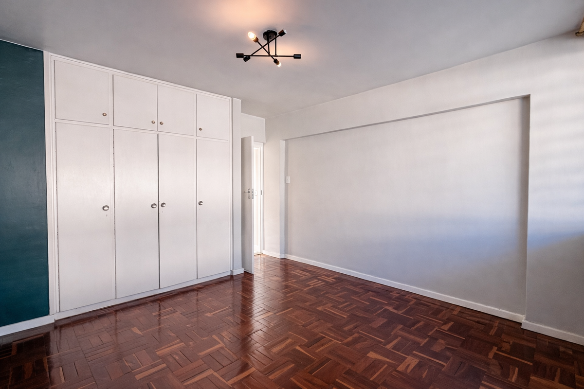 A bright, empty bedroom featuring polished parquet flooring, ample built-in cupboard storage, and a contemporary ceiling light fixture.