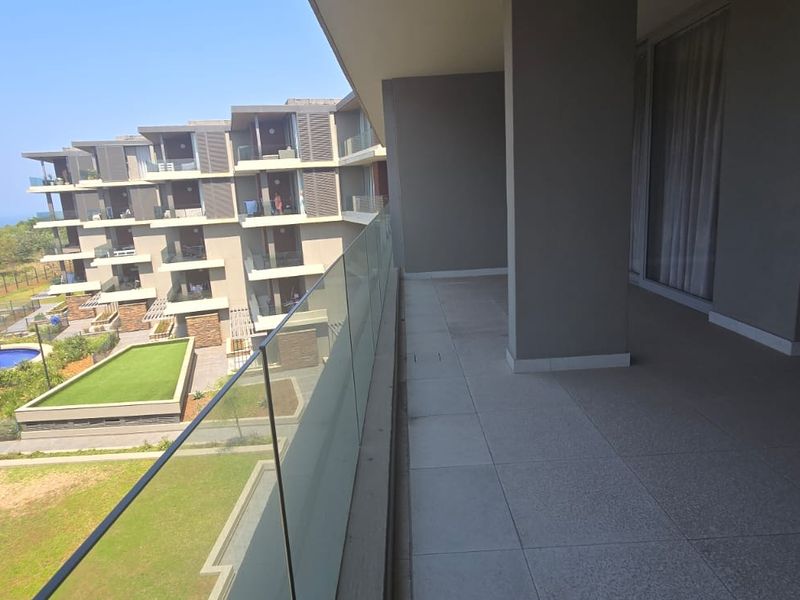 A spacious covered balcony featuring modern glass balustrades, tiled flooring, and views overlooking the apartment complex grounds and swimming pool area.