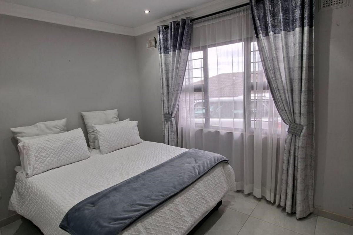 A well-lit bedroom featuring tiled flooring, neutral wall tones, and a window fitted with security bars and curtains.