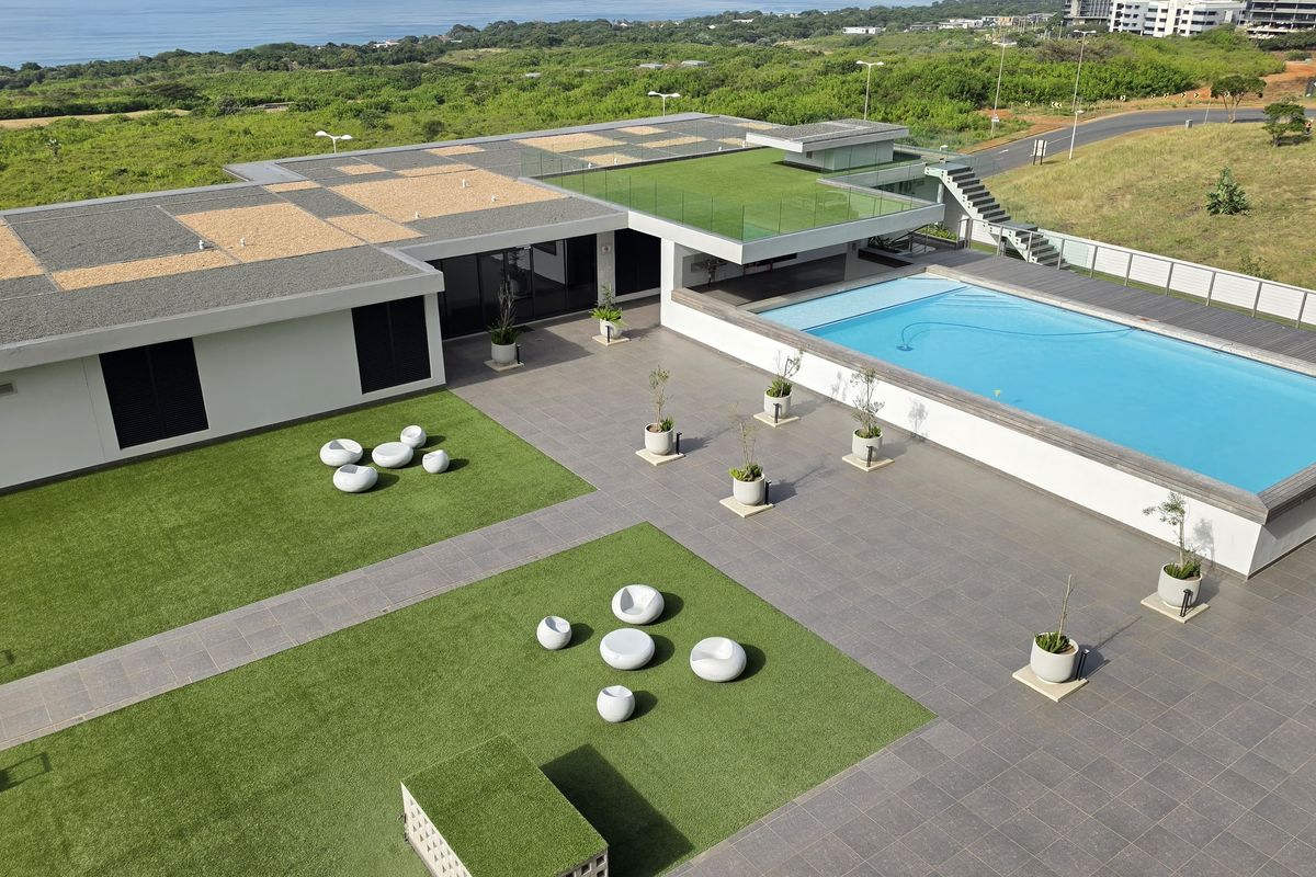 Aerial view of a modern apartment complex featuring a large swimming pool, paved terrace, landscaped artificial grass areas, and a rooftop garden.